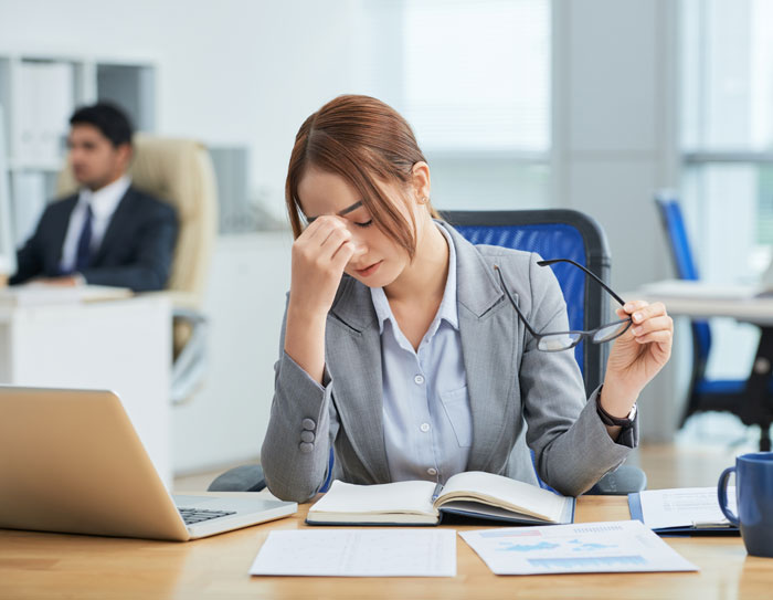 A woman at a desk with her hand on her forehead, looking stressed, holding glasses. This image can be used for boss stories that make you quit your job.
