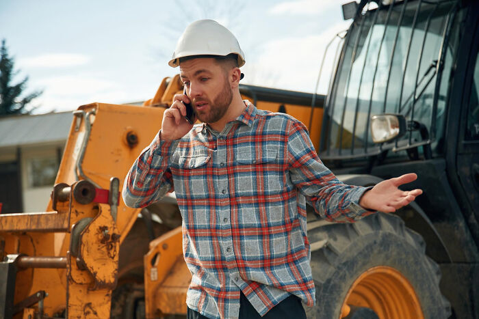 A construction worker in a hard hat talking on his phone with an excavator in the background, a busy environment where pocket dial stories could happen.