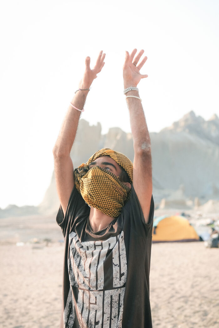A man with a patterned headscarf covering his face raises his arms to the sky in a sandy desert of the Strait of Hormuz.