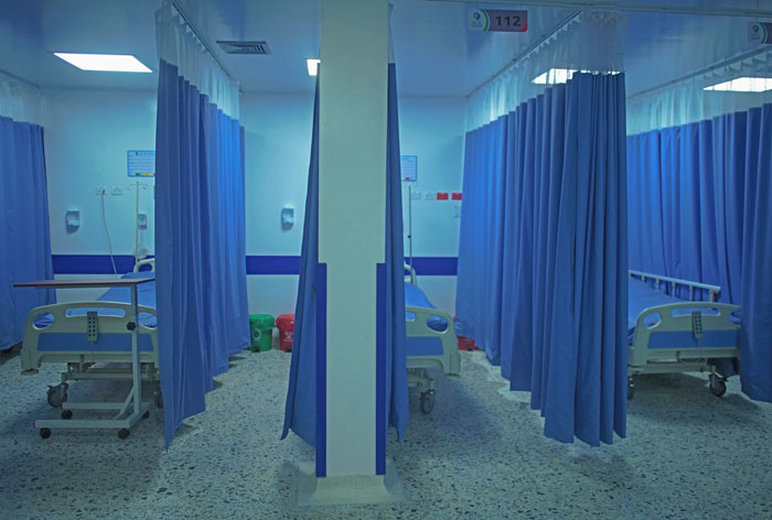 An empty hospital room with beds separated by blue curtains, revealing a hidden side of hospitals.