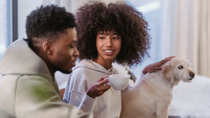 A smiling woman holding a cup, sitting next to a man and petting a golden retriever. Dating scenarios.