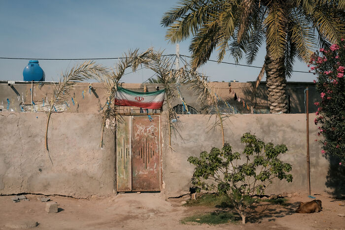 A flag hangs over a weathered gate and stone wall, with palm trees and a blue water tank in the Strait of Hormuz.