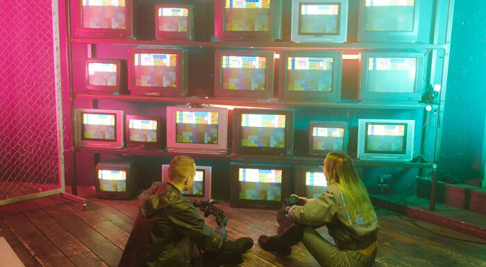 Two people sit on a wooden floor, facing a wall of retro TVs displaying static, exploring their music taste.