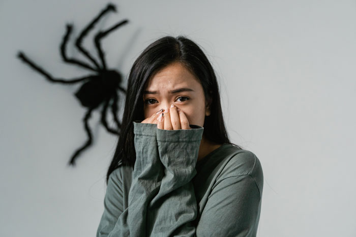 A woman panics, covering her mouth and nose, with a large spider shadow on the wall behind her, highlighting her fear.