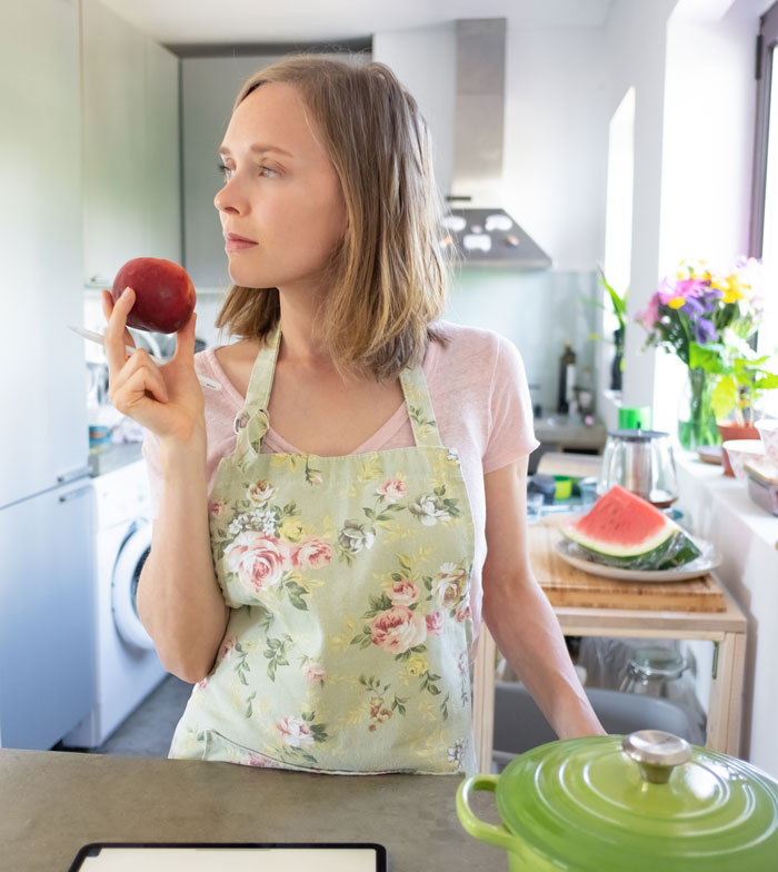 A woman in an apron, contemplating while holding a fruit, representing the challenges after having a baby.