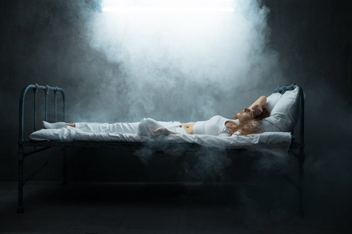 A woman lying on an old bed in a mysterious, smoke-filled room with a bright light overhead, depicting creepy and mysterious things.