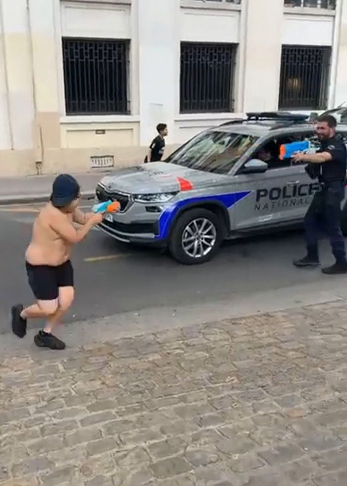 A 14-year-old boy in shorts and no shirt playfully aims a water toy at a police officer by a police car. A 14-year-old boy in shorts and no shirt playfully aims a water toy at a police officer by a police car.
