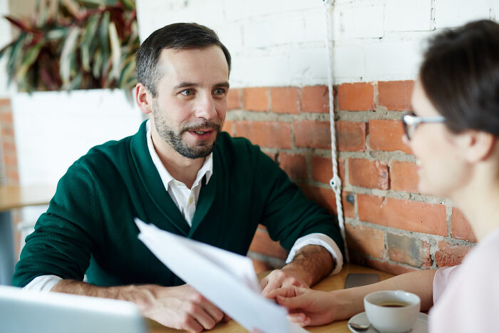 A man in a green cardigan talks to a woman in a cafe, suggesting a conversation about dumb reasons people got fired.