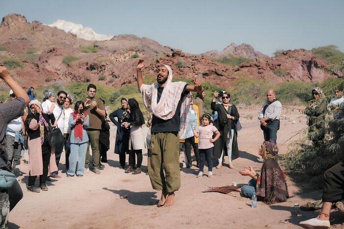 A man with arms raised dances in a dusty clearing surrounded by onlookers and red rock mountains in the Strait of Hormuz.