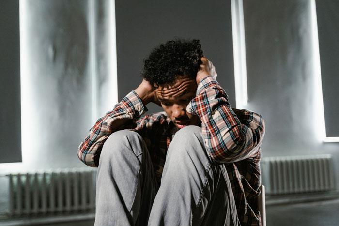 A person with curly hair, wearing a plaid shirt and light pants, sits with their hands covering their head, showing fear.