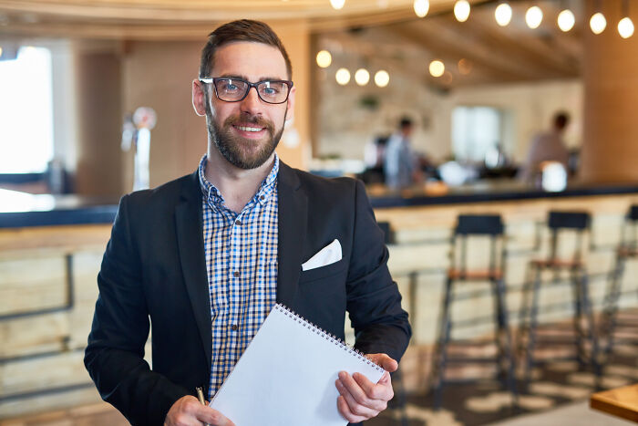 A smiling man in a suit jacket holding a notebook, depicting jobs guys have that make women swipe left.