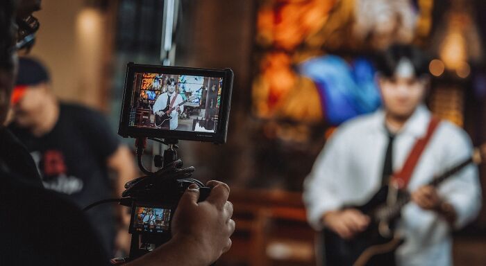 A person filming a musician playing an electric guitar, with the live recording visible on the camera screen, relating to music taste.