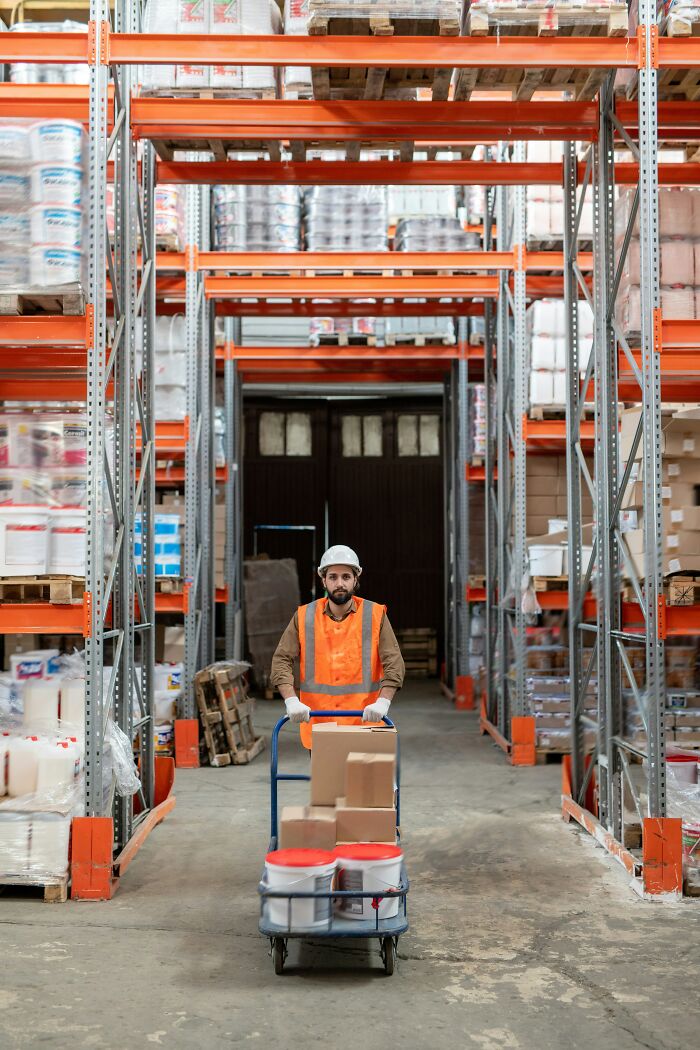 A warehouse worker smiles while pushing a dolly with boxes and buckets down an aisle, a common reason people got fired from their jobs.