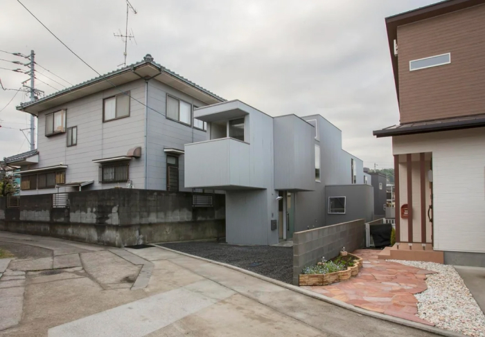 A contemporary modern architecture design house with gray siding, standing between two traditional homes on a cloudy day.