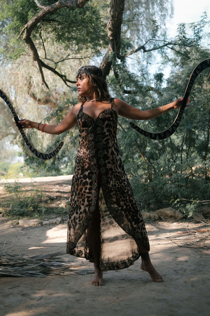 A woman in a leopard print dress, holding two curved objects, performing in a natural setting near the Strait of Hormuz.