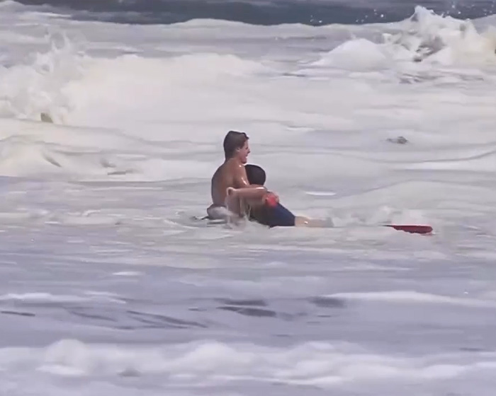 A 16-year-old lifeguard performs a heroic ocean rescue, supporting a person amidst breaking waves. A 16-year-old lifeguard performs a heroic ocean rescue, supporting a person amidst breaking waves.