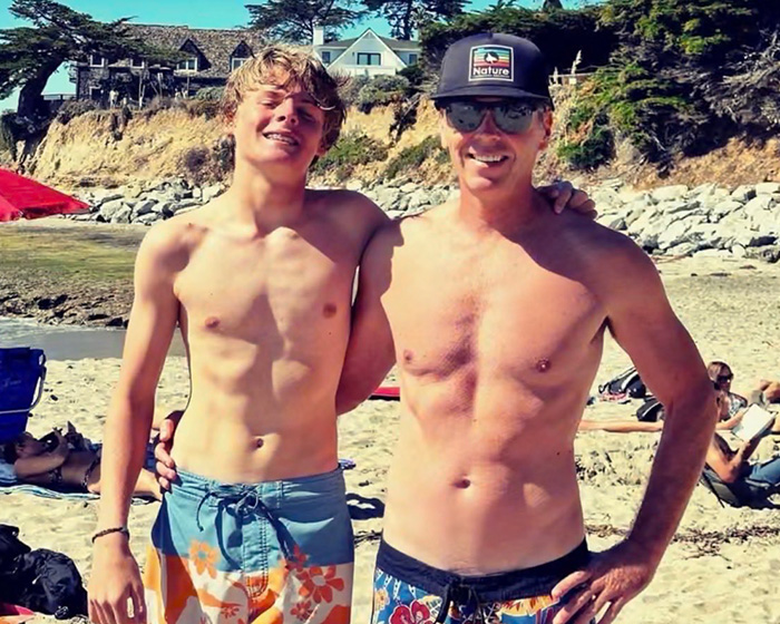 A 16-year-old lifeguard and an adult standing shirtless on a sunny beach after an ocean rescue. A 16-year-old lifeguard and an adult standing shirtless on a sunny beach after an ocean rescue.