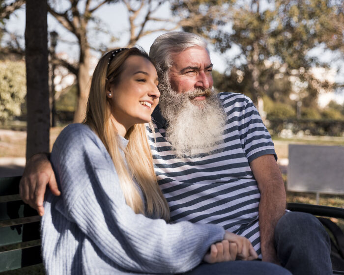 A happy young woman and an older man with a long white beard smiling on a park bench, showing age-gap relationships.