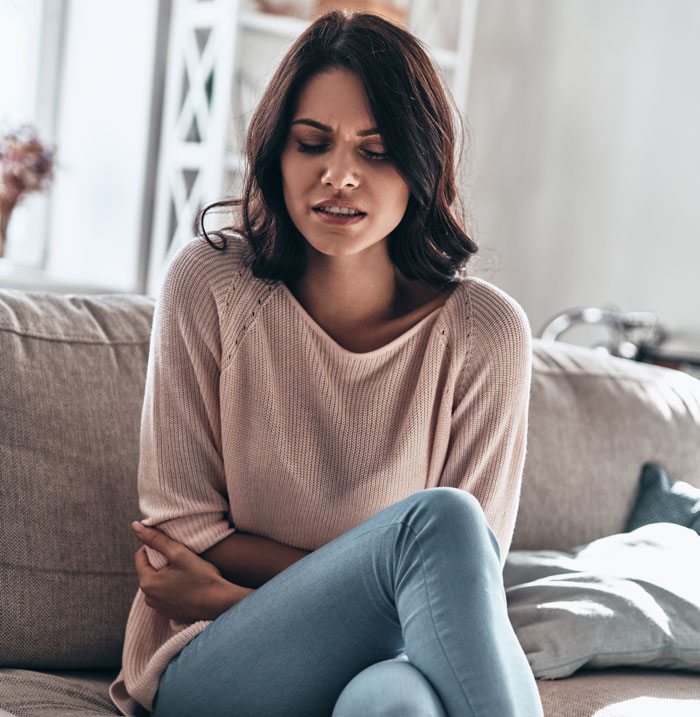 A woman sitting on a couch, holding her stomach in discomfort. A hard truth for women.