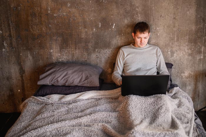 A man sitting in bed, covered by a blanket, using a laptop, representing unhygienic habits.