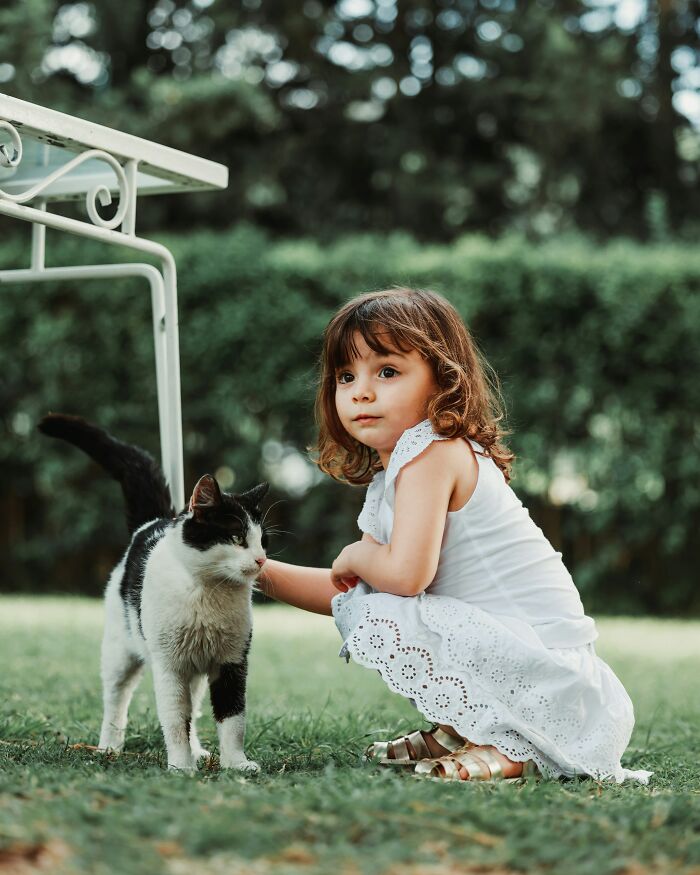 A young girl in a white dress squatting to pet a black and white cat outdoors, showing kids interacting with the world.