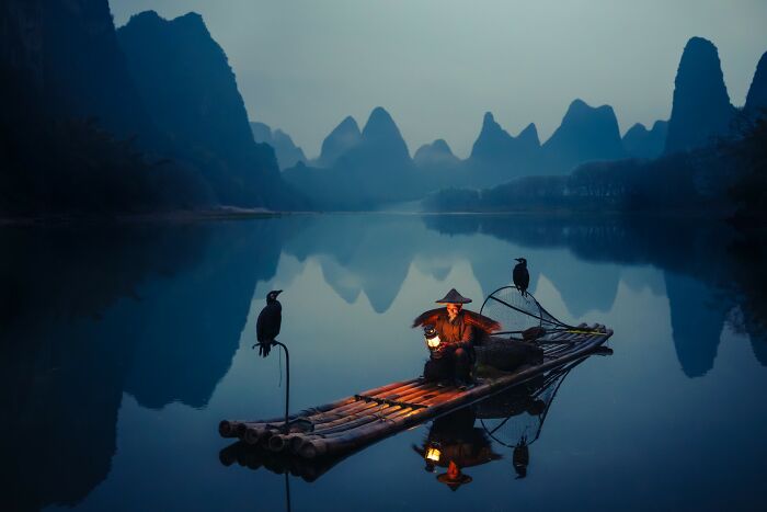 Remarkable color photo of a fisherman on a raft with cormorants on a calm lake with mountains.