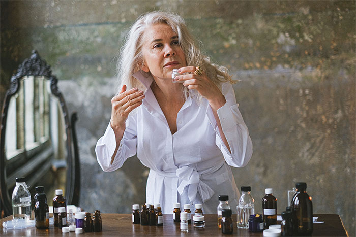 A woman with grey hair in a white shirt, surrounded by small bottles of witching potions, looking closely at one.
