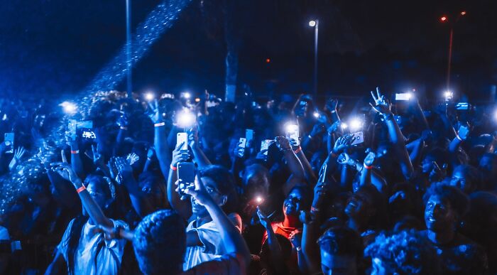 Concert crowd holding up phones with flashlights on, illuminated by blue stage lights, showcasing diverse music taste.