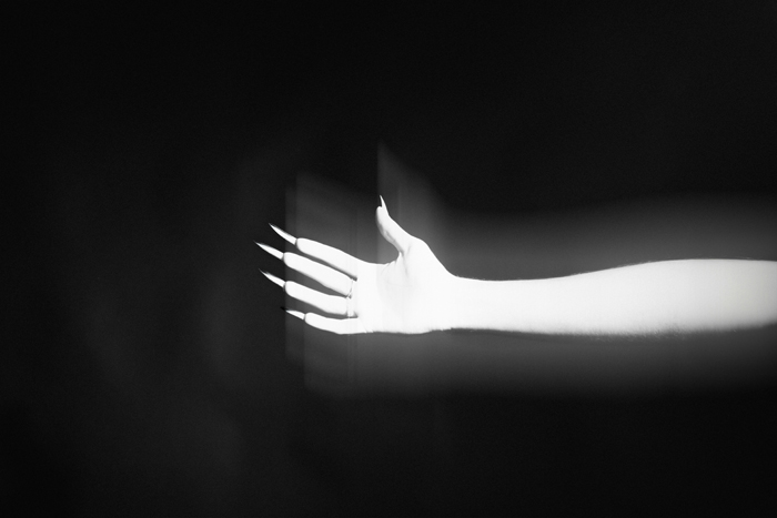 A pale, elongated hand with long, sharp nails reaching out from the darkness, a creepy and mysterious image.