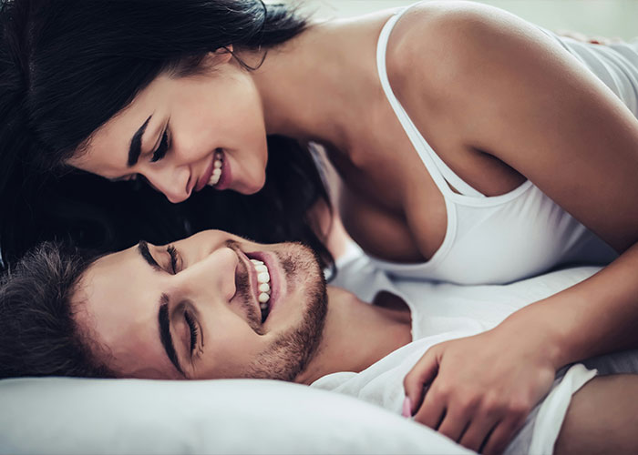 A happy couple smiling in bed, showcasing an intimate position like Queening. A happy couple smiling in bed, showcasing an intimate position like Queening.