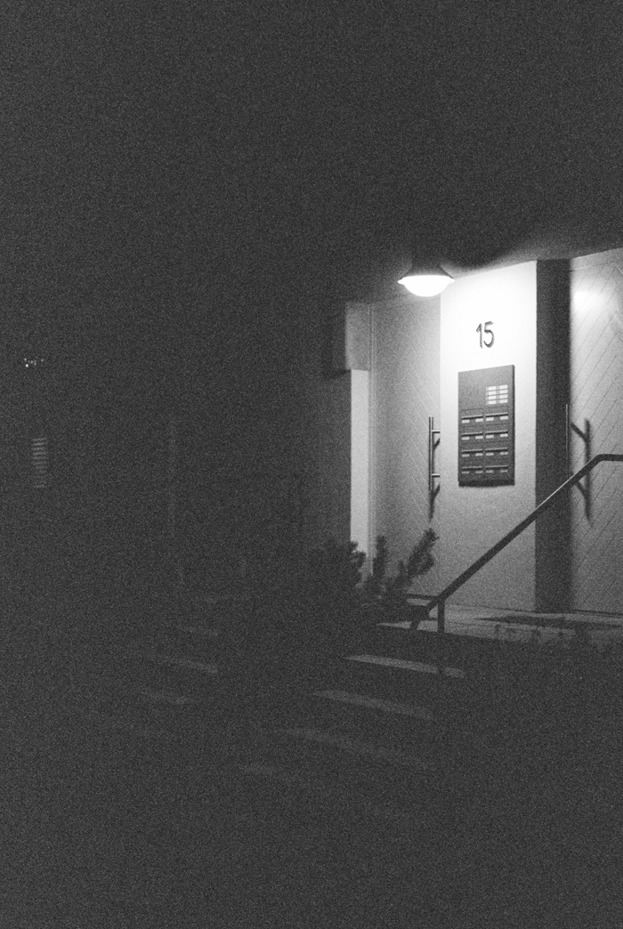A dark, grainy image of an illuminated apartment entrance at night, number 15, adding to the creepy and mysterious mood.