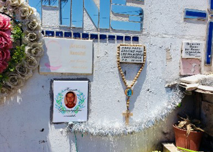 Wall adorned with a rosary, a photo of the disappeared man, and memorial plaques marking his horrific fate.