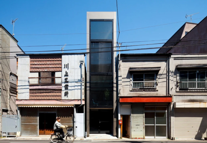 A narrow, modern architecture design building stands tall between two traditional Japanese homes on a sunny day.