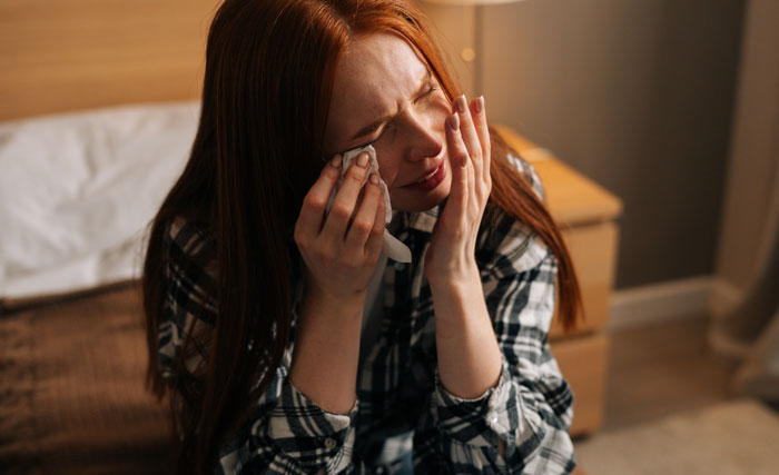 A red-haired woman with eyes closed, crying while holding a tissue, conveying hard truths experienced by women.