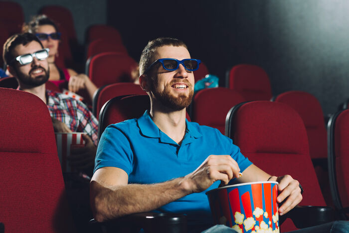 Man wearing 3D glasses and eating popcorn at the cinema, potentially experiencing pocket dial stories aftermath.