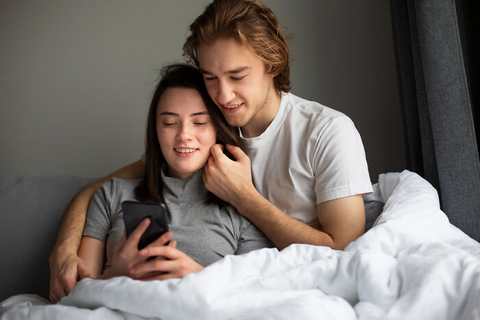 Couple in bed looking at a phone together, possibly sharing or reacting to pocket dial stories.