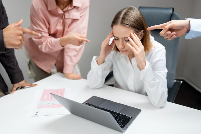 A stressed businesswoman is surrounded by colleagues pointing fingers, illustrating dumb reasons people got fired.