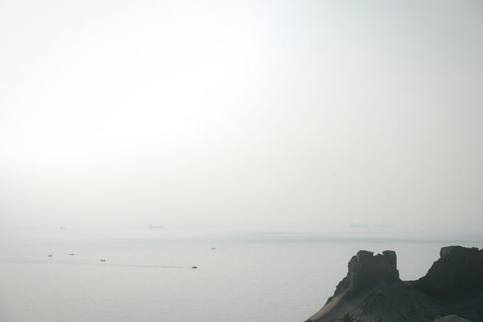 A wide shot of the hazy Strait of Hormuz, with distant ships on the horizon and a rugged coastline in the foreground.
