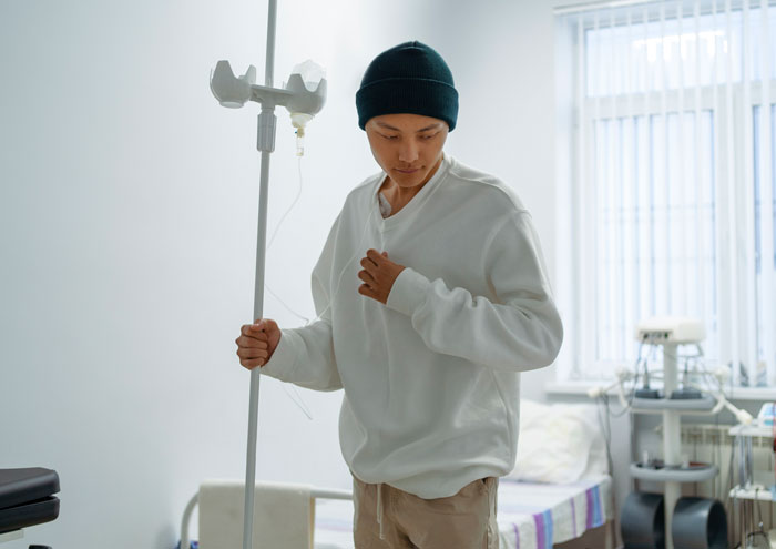 A person in a hospital room, wearing a beanie and an IV, walking with an IV stand, reflecting hard truths.