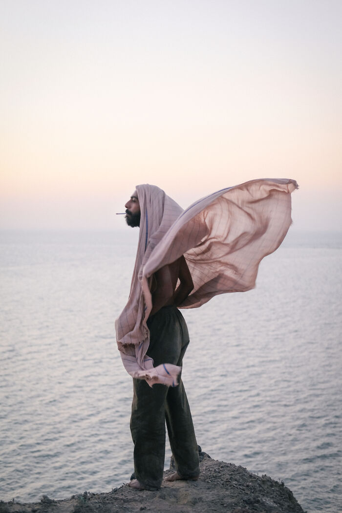 A man with a head covering standing on a cliff overlooking the Strait of Hormuz, with the fabric blowing in the wind.