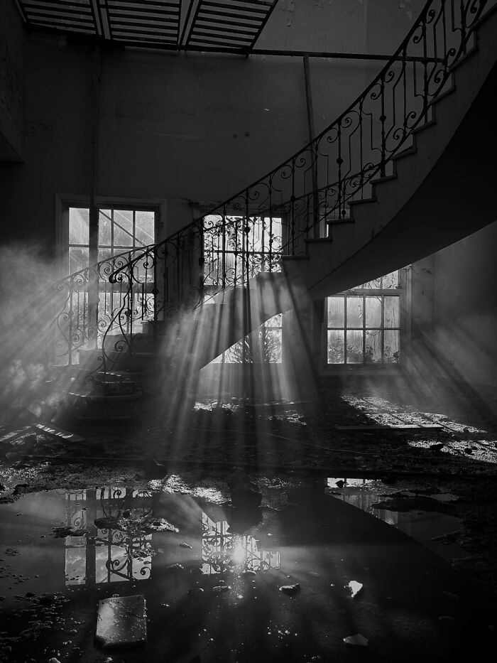 Dramatic light rays illuminate a dark, abandoned room with a staircase and puddles, an extraordinary iPhone Photography Awards photo.