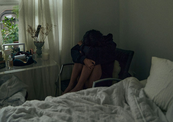 A woman in a dark room, sitting on a chair, hugging her knees to her chest, illustrating hard truths learned.
