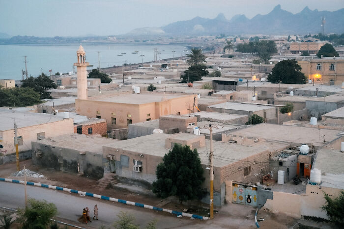 An aerial view of a coastal town by the Strait of Hormuz, featuring a mosque, houses, and mountains in the distance.