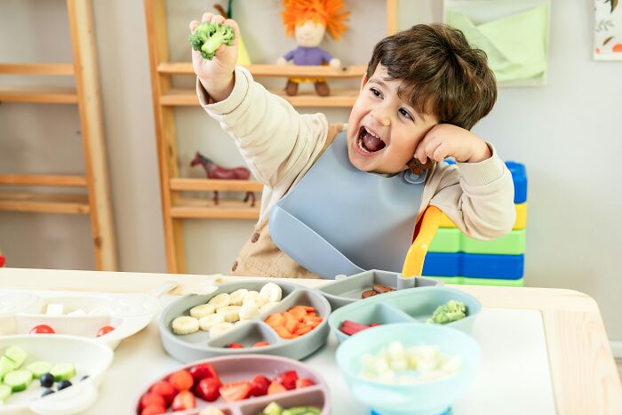 A toddler in a bib excitedly holding up a piece of broccoli, surrounded by various healthy foods, reflecting kids exploration.