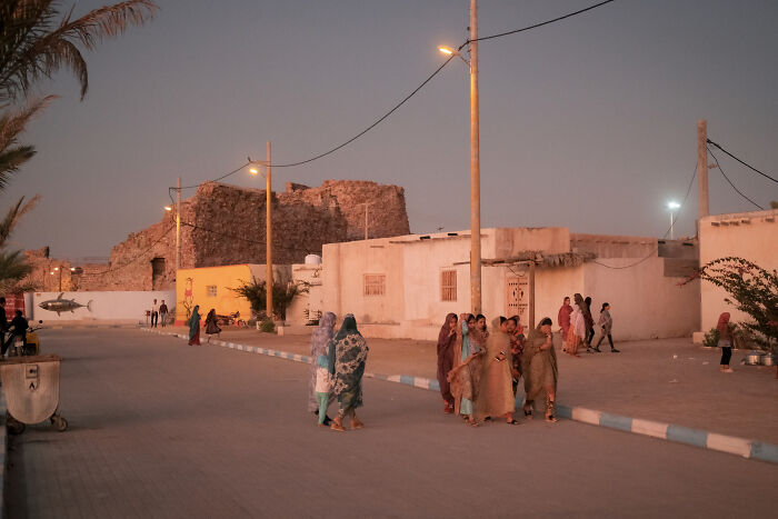 People gather on a street in a town near the Strait of Hormuz, with traditional architecture and old walls in the background.