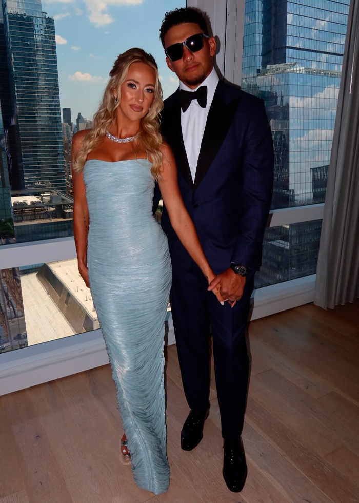 A couple dressed in formal attire stands by a window with a city view. They are celebrities at Taylor Swift's wedding.
