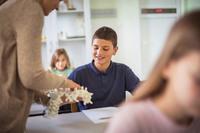 A boy smiling at a teacher in a classroom holding a model, representing kids asking questions and learning.