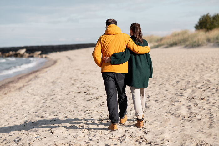 An age-gap couple walking arm-in-arm on a sandy beach, showcasing their relationship together.