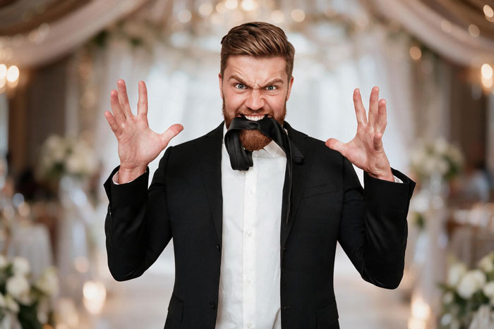 An angry groom in a suit bites his tie, standing in a wedding venue, before his big day. An angry groom in a suit bites his tie, standing in a wedding venue, before his big day.