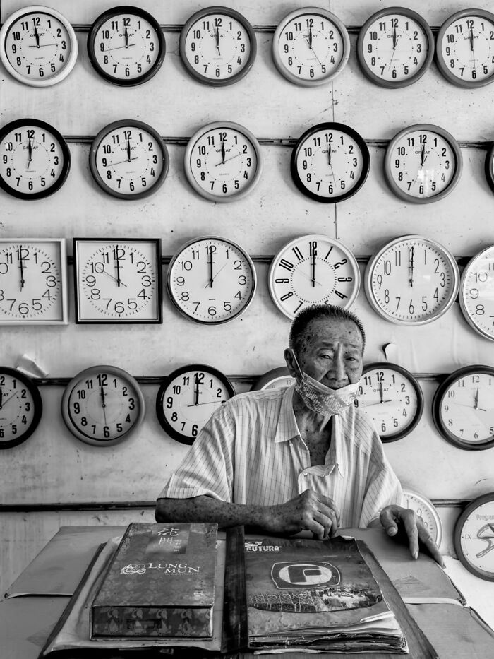 An elderly man wearing a face mask sits in front of a wall of clocks, an extraordinary winning photo from the iPhone Photography Awards.